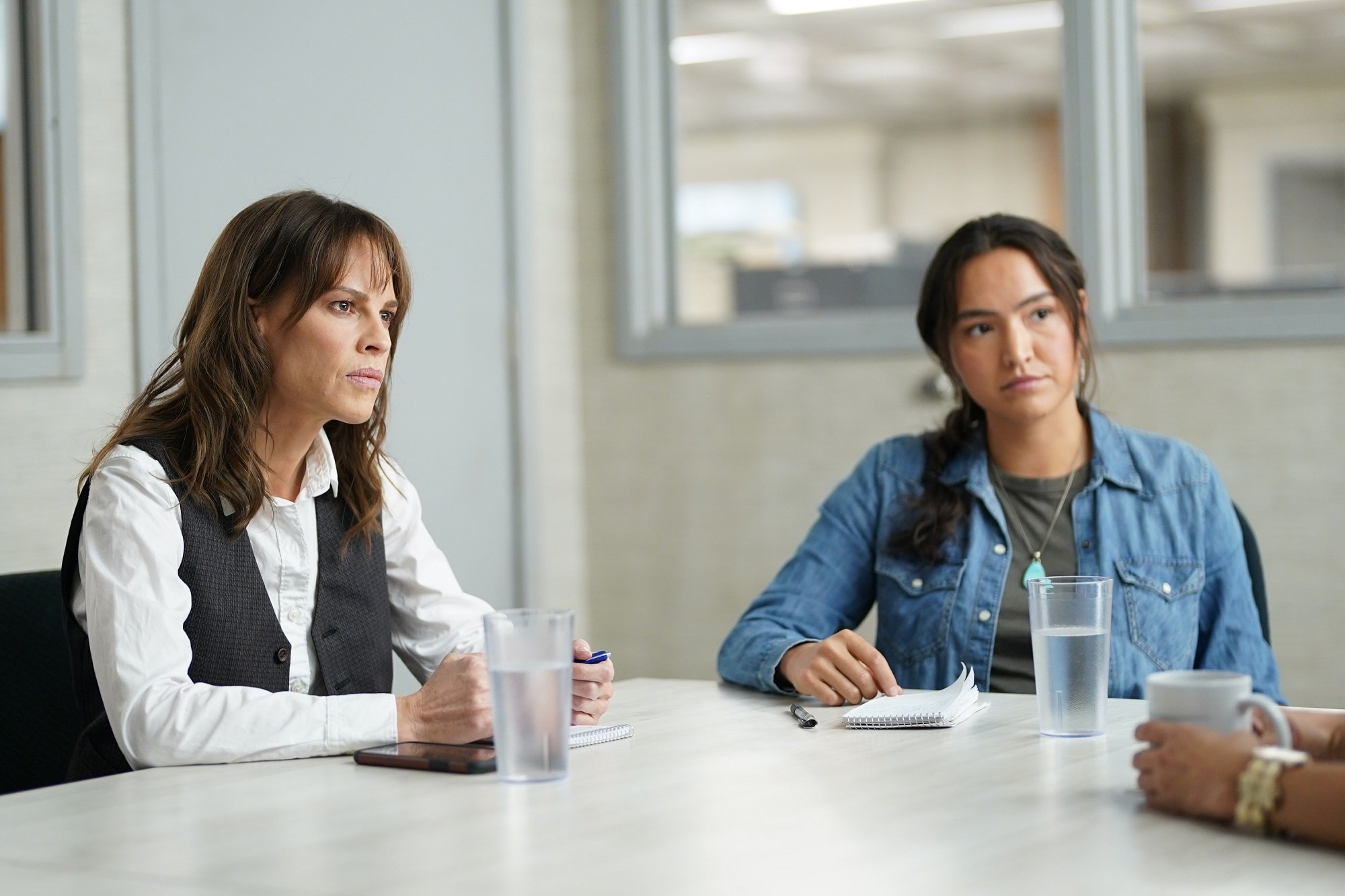 'Alaska Daily' cast members Hilary Swank and Grace Dove sit at a table together intently listening to someone