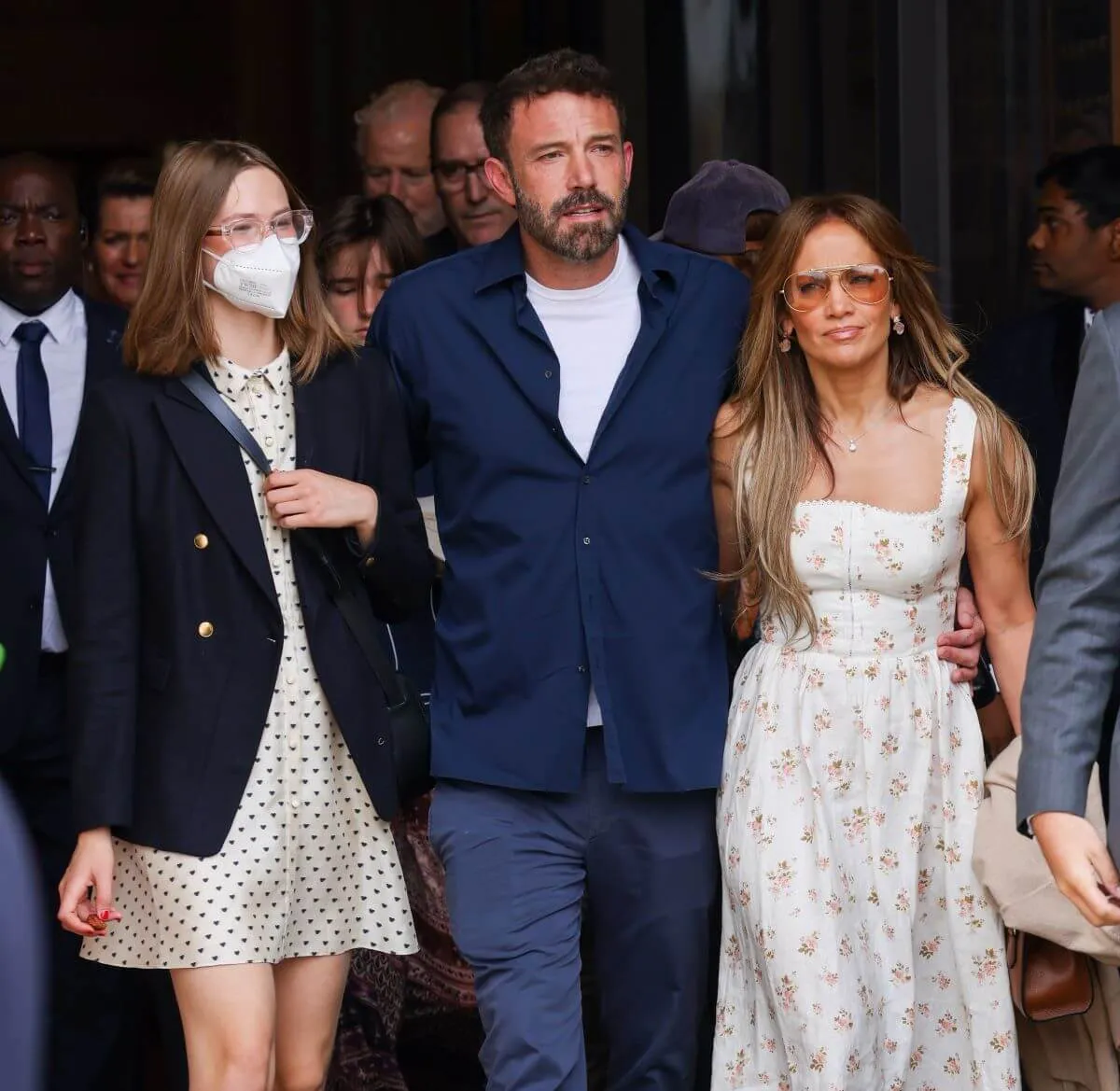 Violet Affleck, Ben Affleck, and Jennifer Lopez walk together. Violet Affleck wears a dress, glasses, and a mask. Ben Affleck wears jeans and a blue shirt. Jennifer Lopez wears sunglasses and a dress.