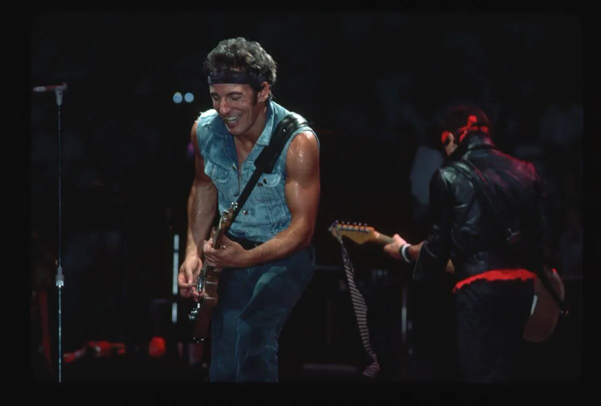 Bruce Springsteen wears a vest and a bandana and plays guitar.