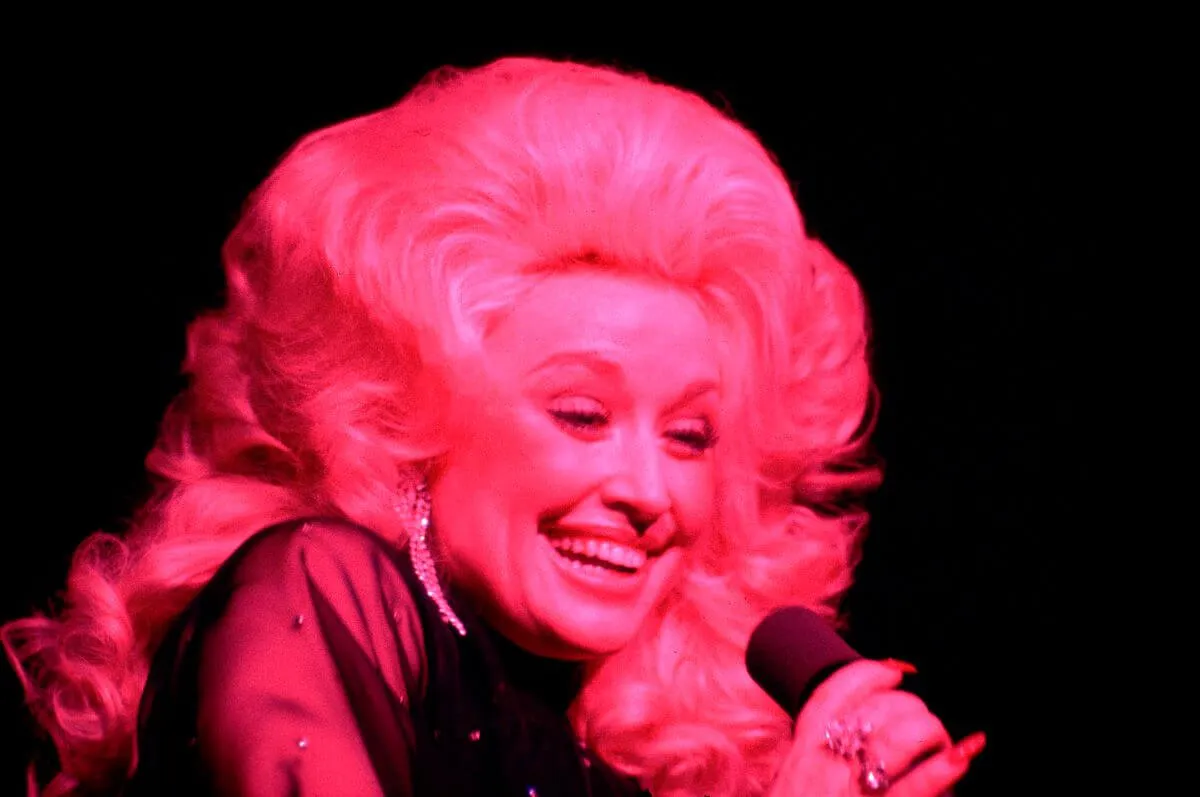 Dolly Parton smiles and holds a microphone. She is lit by pink light.