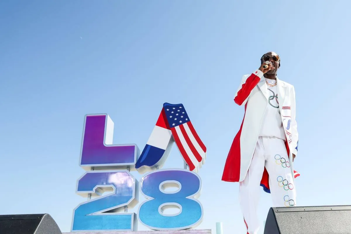 Snoop Dogg holds a microphone and stands in front of a sign for LA28. He wears a red and white coat.